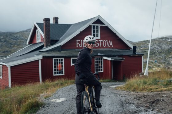 Sabina posing on her gravel bike standing before a red Fjellstova on the Rallarvegen